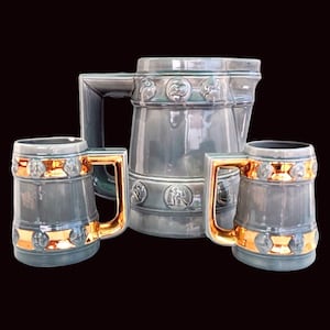May include: A set of three gray ceramic mugs with gold accents. The mugs have a pitcher-like shape and feature embossed designs. The mugs are perfect for serving beverages.