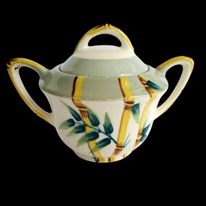 Puede incluir: Un azucarero de cerámica vintage con tapa. El cuenco es blanco con tallos de bambú amarillos y marrones y hojas verdes. La tapa es de color verde claro con un asa amarilla. El cuenco tiene dos asas.