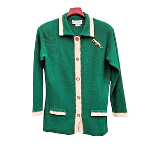 May include: A long-sleeve emerald green knit cardigan featuring a cream-colored collar, button-down front, and matching cream trim on the cuffs and two front pockets. A gold-toned metal panther brooch is pinned to the upper left chest area.