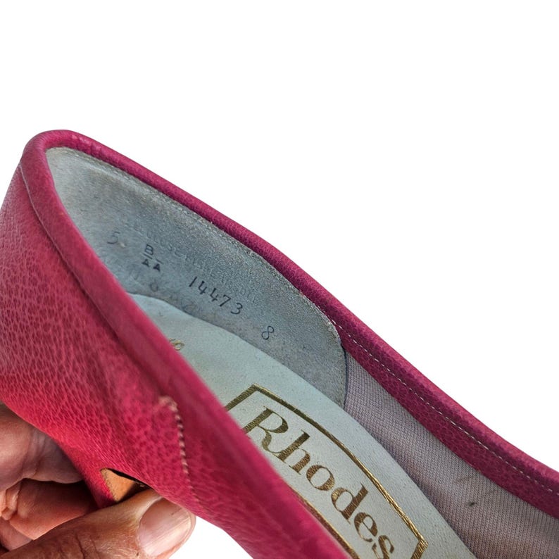 May include: Close-up of a bright pink Rhodes brand shoe. The shoe's interior is visible, showing the size 8 and other markings. The shoe has a textured surface and a stitched trim.