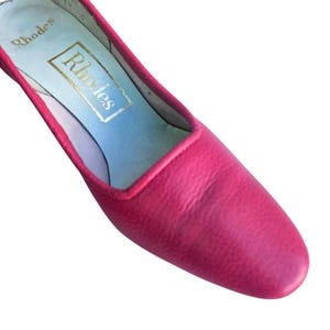 May include: A close-up of a pink leather shoe with a pointed toe. The shoe has a light blue insole with the gold text "Rhodes". The shoe's trim is a darker shade of pink.