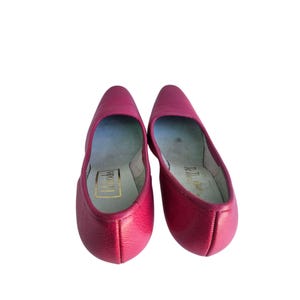 May include: A pair of bright pink leather flats. The shoes have a pointed toe and a low heel. The interior of the shoe is a light blue color with gold lettering that reads "Rhodes". The shoes are set against a white background.