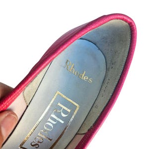 May include: Close-up of a pink leather shoe interior. The shoe's insole is light blue with the gold-colored brand name "Rhodes" printed on it. The shoe's size is 8.