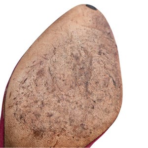 May include: Close-up of the worn sole of a shoe. The sole is a light brown color with dark brown and black markings, showing signs of wear and use. The shoe's edge is a deep red color.