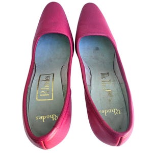 May include: A pair of bright pink leather flats. The shoes have pointed toes and a low heel. The insoles are light blue with gold lettering that reads "Rhodes" and "Mr. Lea".