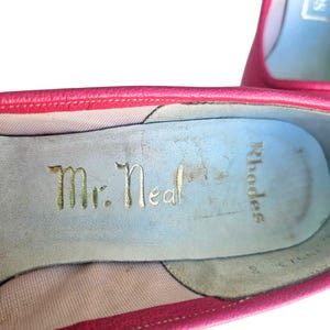 May include: Close-up of a pair of pink leather shoes. The interior of the shoe features the gold text "Mr. Neal" and "Rhodes" on a light blue insole. The shoe's edges are a darker pink.