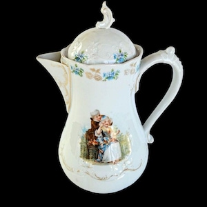 Puede incluir: Cafetera de porcelana blanca con tapa, asa y boquilla decorativas. La cafetera está adornada con motivos florales en azul y dorado, y una escena pintada de dos niños. Una pieza clásica para servir.