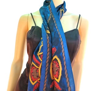 Puede incluir: Una bufanda larga azul con un patrón repetitivo de bordes de cuerda marrón, formas de escudo amarillas y motivos de flor de lis rojos. La bufanda está colocada alrededor del cuello, mostrando una paleta de colores vibrantes en azul, amarillo y rojo.