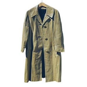 May include: A vintage, light olive green leather coat hangs from a wooden hanger. The coat features a notched collar, button closure, and long sleeves. The coat has a knee-length hemline and visible stitching details.