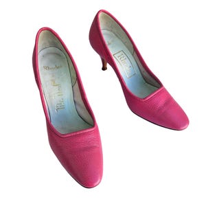 May include: A pair of bright pink, vintage Rhodes heels. The shoes have a low heel and a pointed toe. The interior of the shoe is light blue with the Rhodes logo in gold. The shoes are made of a textured leather material.