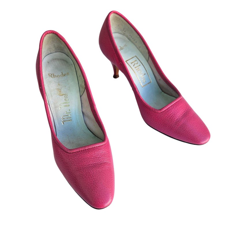 May include: A pair of bright pink, vintage Rhodes heels. The shoes have a low heel and a pointed toe. The interior of the shoe is light blue with the Rhodes logo in gold. The shoes are made of a textured leather material.