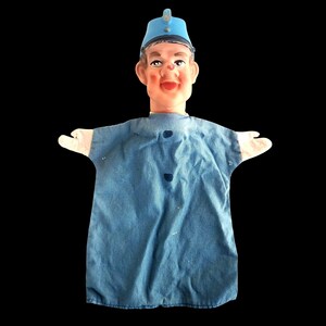 May include: A vintage hand puppet featuring a painted face with rosy cheeks, a blue cap, and a blue denim-like shirt. The puppet's arms are outstretched, with white hands and a black background.
