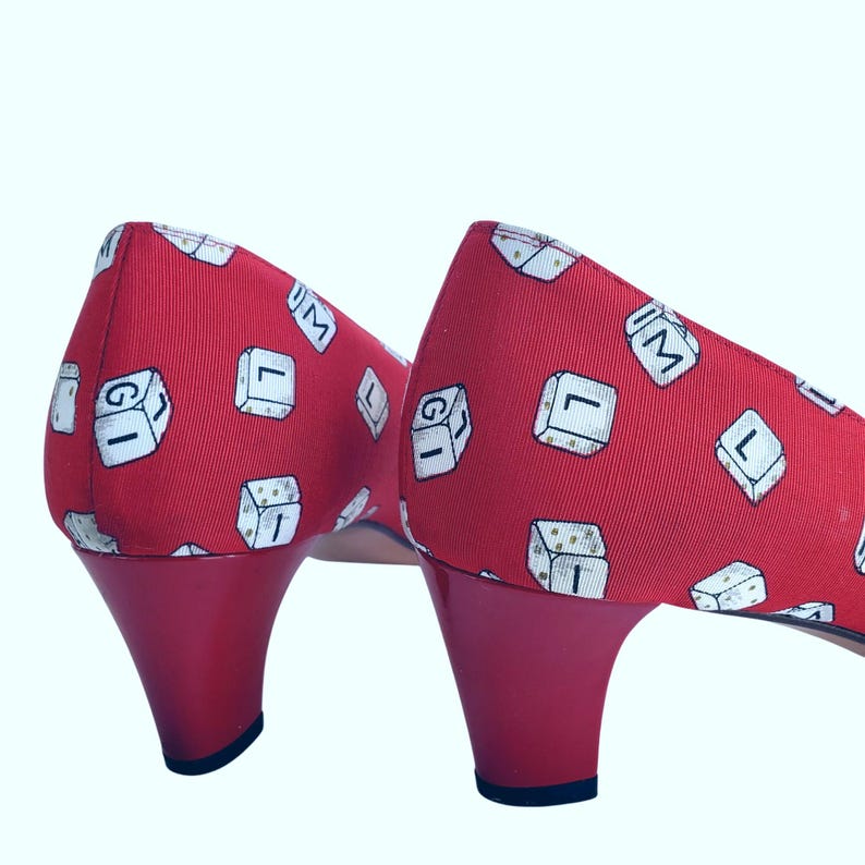 May include: A pair of red heels with a Scrabble tile pattern. The heels have a glossy red heel and a rounded toe. The fabric is red with white tiles featuring letters and numbers. The heels are a low to mid-height.