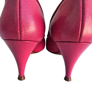 May include: A pair of bright pink high heels with a textured leather appearance. The heels are a stiletto style, with a brown tip. The shoes are set against a white background.