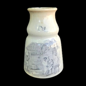 May include: A vintage, off-white ceramic jar with a flared base and a narrow neck. The jar features a detailed illustration of a market scene with the text "LUCKNOW CHUTNEY". The jar has a slightly aged appearance.