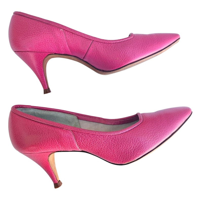 May include: Two pairs of bright pink, pointed-toe pumps with textured leather uppers. The shoes have a low, curved heel and a classic silhouette, perfect for a vintage or retro look.