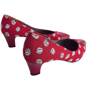 May include: A pair of red heels with a low heel. The shoes have a red fabric upper with a repeating pattern of white keyboard keys. The toe cap is a darker red leather.