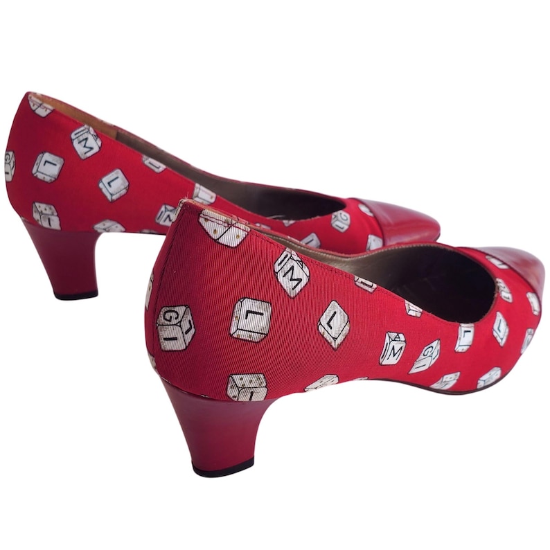 May include: A pair of red heels with a low heel. The shoes have a red fabric upper with a repeating pattern of white keyboard keys. The toe cap is a darker red leather.
