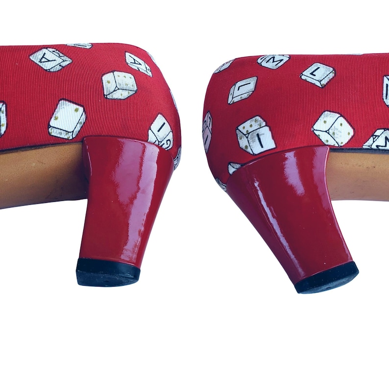 May include: Pair of red heels with a dice pattern on the fabric. The heels have a glossy red finish and a black sole. The shoes are a playful and unique fashion statement.