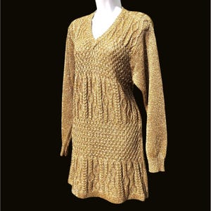 Può includere: Un abito in maglia dorato a maniche lunghe con scollo a V e motivo a maglia strutturata. L'abito ha un corpetto aderente e un orlo svasato. L'abito è esposto su un manichino bianco su sfondo nero.