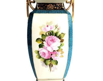 May include: A rectangular porcelain vase with a cream-coloured central panel featuring a bouquet of pink roses and green leaves. The sides are teal with a textured pattern, and the top and handles are gold. The base is black.