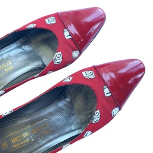 May include: A pair of red heels with a glossy finish on the toe caps. The fabric features a repeating pattern of white dice with numbers and letters. The shoes are from the brand Bruno Magli.