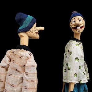May include: Two wooden marionette puppets with painted faces and wearing hats and shirts. One puppet has a long nose and a blue and green striped knit cap. The other puppet has a tooth and a blue knit cap. The background is black.
