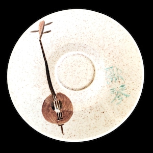 Könnte beinhalten: Eine weiße Keramikuntertasse mit einem gesprenkelten Muster und einer zentralen Vertiefung. Die Untertasse zeigt eine handgemalte Illustration eines Saiteninstruments in Braun und Schwarz. Auf der Untertasse befindet sich auch blauer Text.
