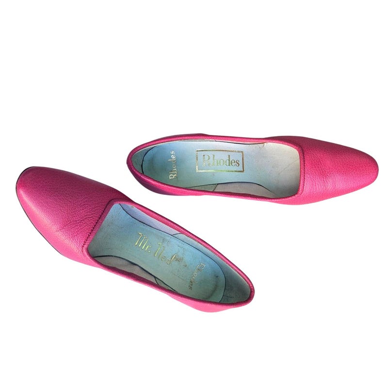 May include: A pair of bright pink leather flats. The shoes have a pointed toe and a low profile. The interior of each shoe has a light blue insole with gold lettering that reads "Rhodes" and "Mr. Ted".