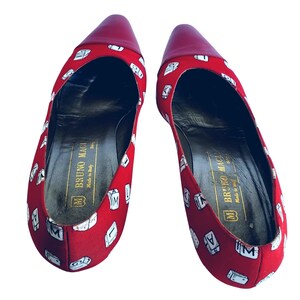May include: A pair of red pointed-toe flats with a repeating white letter design. The toe and heel are a solid red. The interior of the shoe is black with gold text that reads "BRUNO MAGLI Made in Italy".