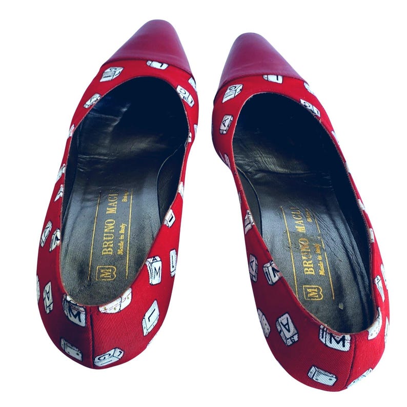 May include: A pair of red pointed-toe flats with a repeating white letter design. The toe and heel are a solid red. The interior of the shoe is black with gold text that reads "BRUNO MAGLI Made in Italy".