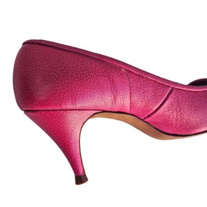 May include: A close-up of a bright pink leather pump with a pointed toe and a slender, curved heel. The shoe has a textured surface and a classic design. The heel is approximately 2 inches (5 cm) high.