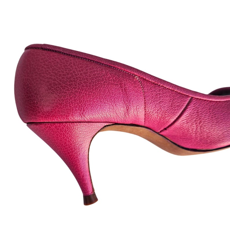 May include: A close-up of a bright pink leather pump with a pointed toe and a slender, curved heel. The shoe has a textured surface and a classic design. The heel is approximately 2 inches (5 cm) high.