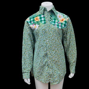 Könnte beinhalten: Grünes Hemd mit Blumenmuster. Das Hemd hat ein Patchwork-Design auf der Brust mit Gänseblümchen und einem grünen Vichy-Muster. Es hat lange Ärmel und einen klassischen Kragen.