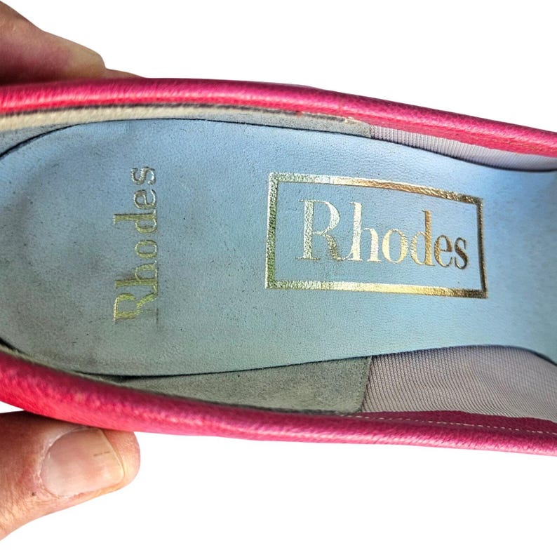 May include: Close-up of a pink shoe interior with the brand name "Rhodes" in gold lettering. The insole is light blue, and the shoe's edge is a vibrant pink. The shoe is held by a hand.