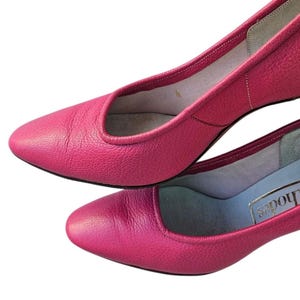 May include: Two pairs of bright pink leather pumps. The shoes have a low heel and a rounded toe. The interior lining is a light color. The shoes are set against a white background.