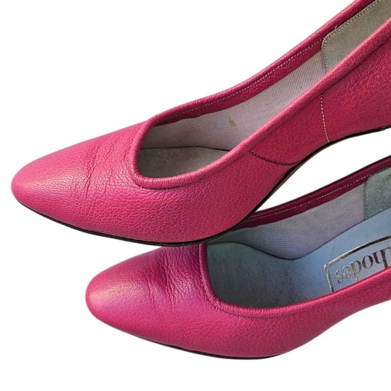 May include: Two pairs of bright pink leather pumps. The shoes have a low heel and a rounded toe. The interior lining is a light color. The shoes are set against a white background.