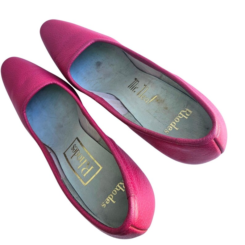 May include: A pair of bright pink leather flats. The shoes have pointed toes and a low heel. The insoles are light blue with gold lettering that reads "Rhodes" and "Mr. Neal".