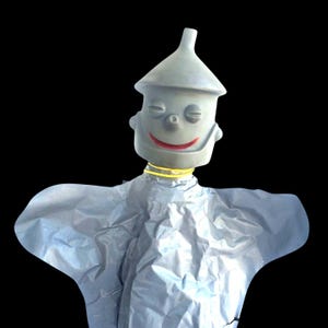 Puede incluir: Una figura gris del Hombre de Hojalata con un sombrero en forma de cono y una cara sonriente. La figura tiene un collar amarillo y un cuerpo plateado, sobre un fondo negro. La figura es un personaje de El Mago de Oz.