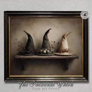 May include: Framed art print featuring three witch hats on a wooden shelf. The hats are black, gray, and cream-colored, adorned with floral and botanical accents. The artwork is signed "The Decadent Witch Fine Art Prints."