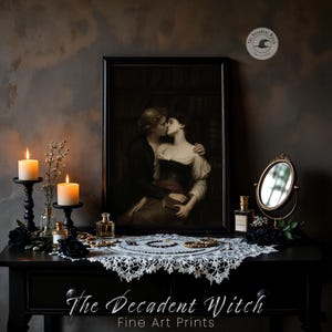 May include: A black table displays a framed artwork of a couple kissing, flanked by lit candles in black holders, perfume bottles, and a small mirror. A white lace doily and jewelry adorn the table. The text "The Decadent Witch Fine Art Prints" is visible.