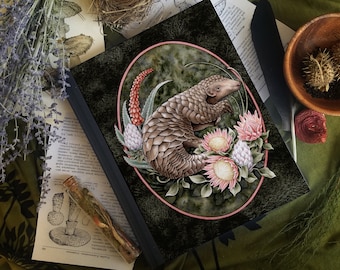 May include: A hardcover journal featuring a detailed illustration of a pangolin surrounded by flowers and foliage within a pink oval frame. The cover has a dark green background and a black border.
