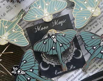 May include: Five enamel pins featuring a green and gold luna moth design. The pins are arranged on a surface with a book and a piece of bark. The pins are all different sizes and have a glossy finish. The text "Moon Magic" and "Enamel Pin" are visible on the pins.