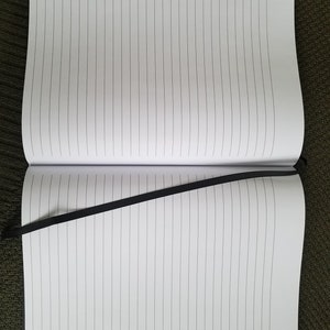 Puede incluir: Un cuaderno negro con l&iacute;neas y un marcador de cinta negro. El cuaderno est&aacute; abierto en una p&aacute;gina en blanco.