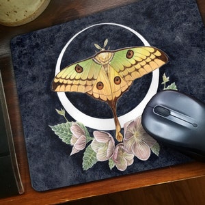 May include: A mouse mat featuring a watercolour illustration of a green and yellow luna moth with brown markings, perched on a white crescent moon. The moth is surrounded by pink and white flowers with green leaves on a dark blue background.