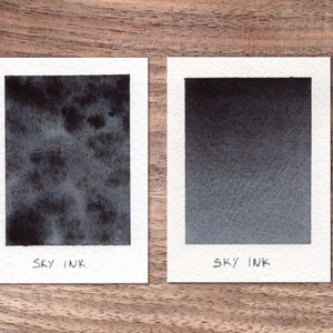 Könnte beinhalten: Zwei quadratische Aquarellmalereien auf weißem Papier, jede mit einer dunkelblau-grauen Lasur. Das linke Gemälde hat ein strukturiertes, gesprenkeltes Aussehen, während das rechte einen glatten Farbverlauf aufweist. Beide sind mit "SKY INK" beschriftet.