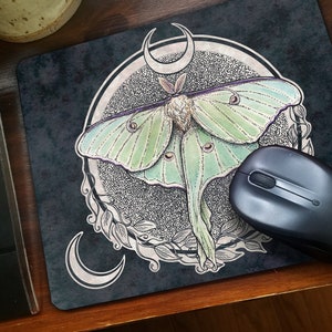 May include: A black mouse pad with a green and white luna moth illustration. The moth is surrounded by a circle with a crescent moon design. The mouse pad is on a wooden surface.