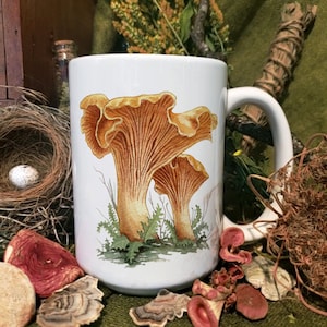 Puede incluir: Taza de cerámica blanca con una ilustración colorida de dos rebozuelos. Los hongos se representan de forma realista con tonos marrones y amarillos. La taza tiene un asa blanca.