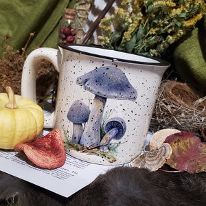 May include: White ceramic mug with a black rim and handle. The mug features a colorful illustration of three mushrooms with black speckles on a white background.