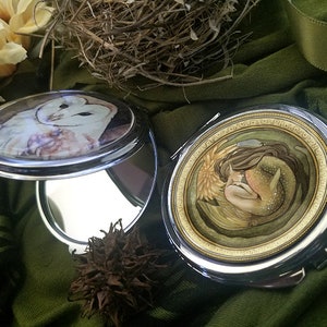 Puede incluir: Dos espejos compactos plateados con diseños diferentes. Un espejo presenta un búho blanco con plumas marrones, el otro presenta una sirena con cabello largo marrón y una cola dorada.